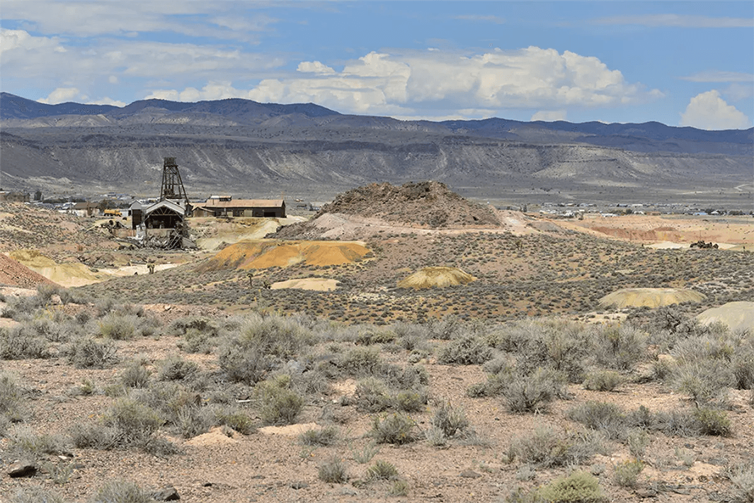 A desert landscape with shrubs, old wooden structures and mountains under a partly cloudy sky.