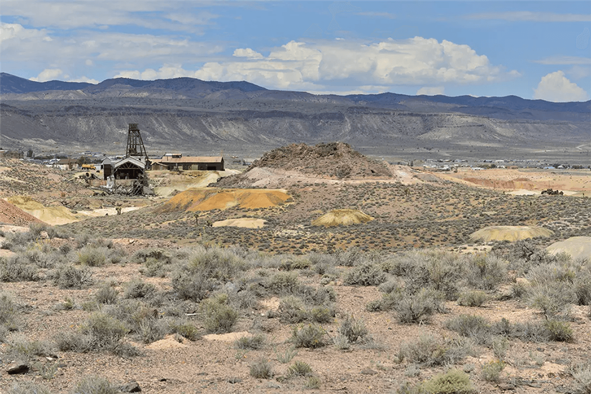 A desert landscape with shrubs, old wooden structures and mountains under a partly cloudy sky.