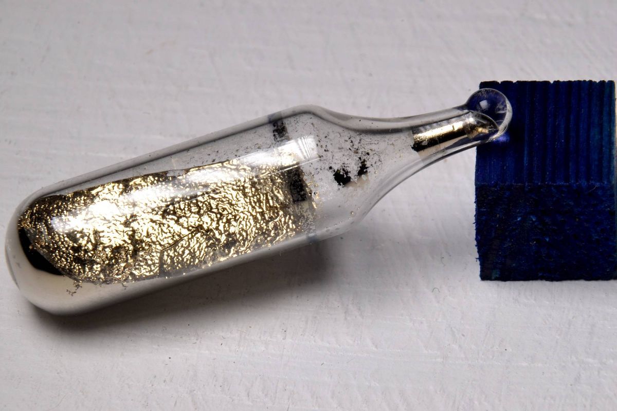 A glass vial containing a cesium metal sample is placed next to a textured blue block on a white surface.