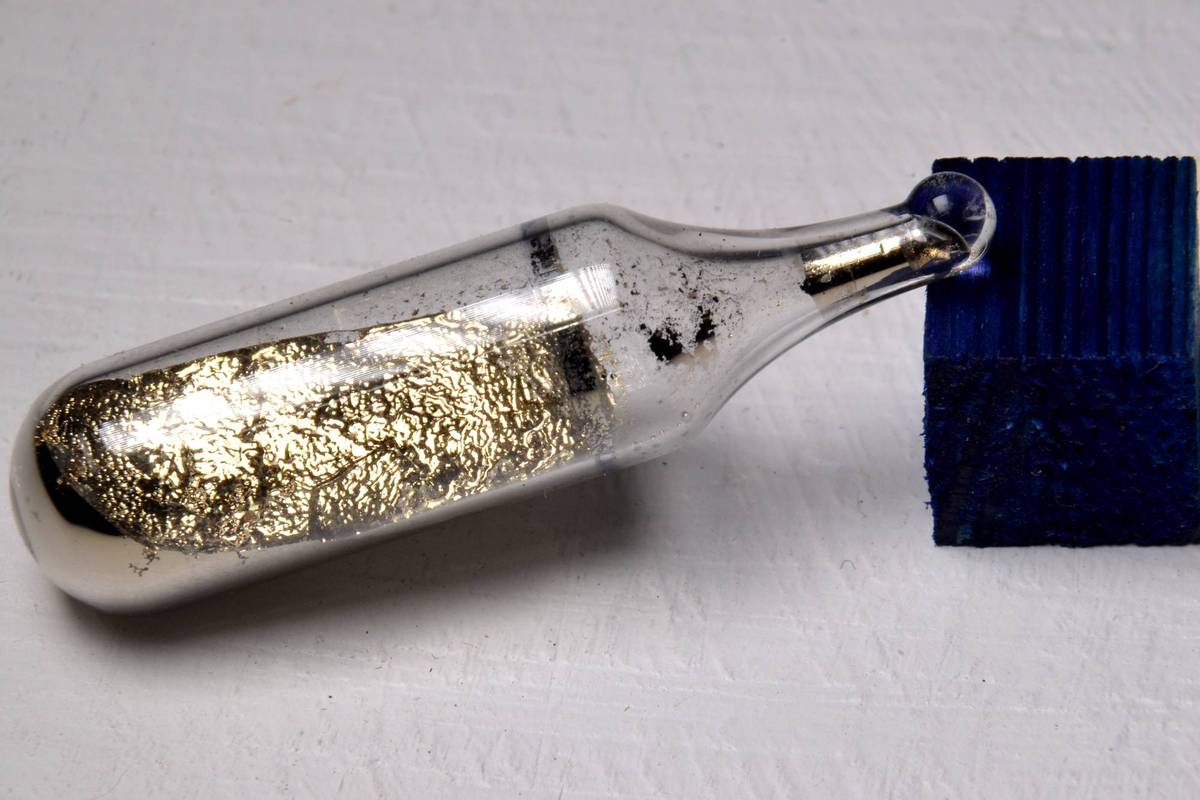 A glass vial containing a cesium metal sample is placed next to a textured blue block on a white surface.
