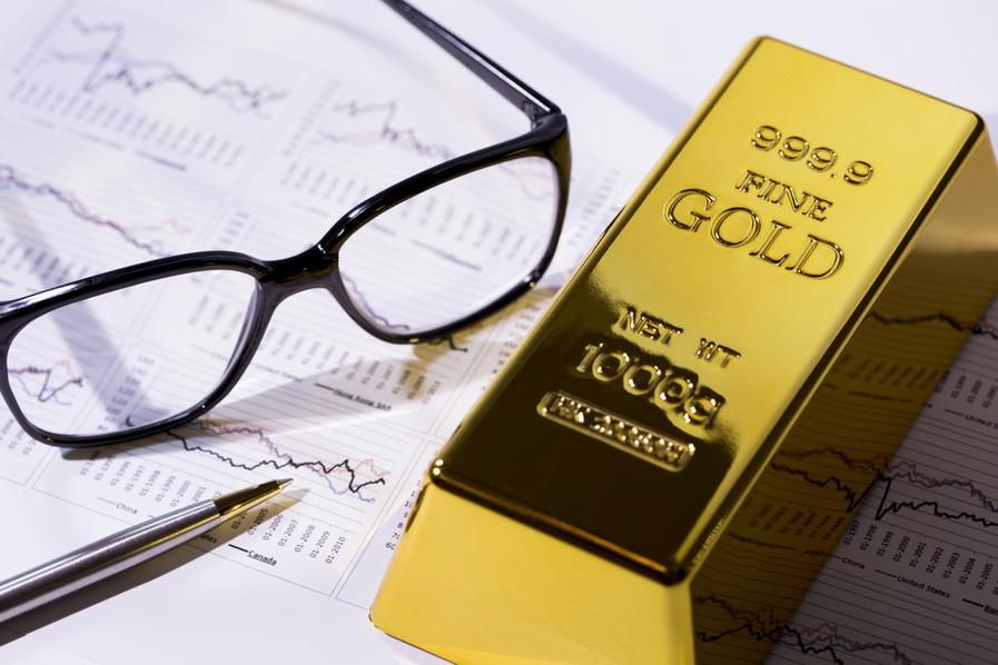a gold bar beside a pair of glasses and pen atop stock charts