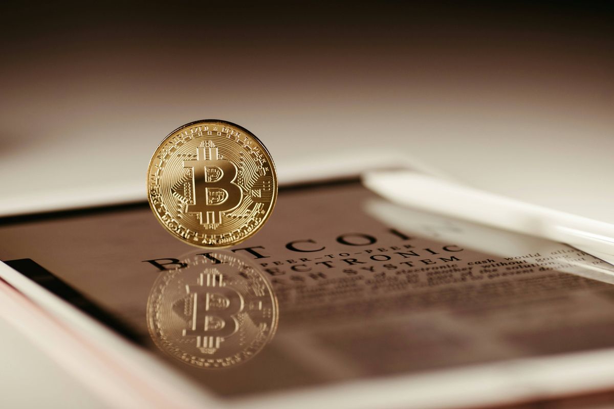 A gold Bitcoin coin over a document page with partial text: BTC, ELECTRONIC SYSTEM, reflected on the surface.