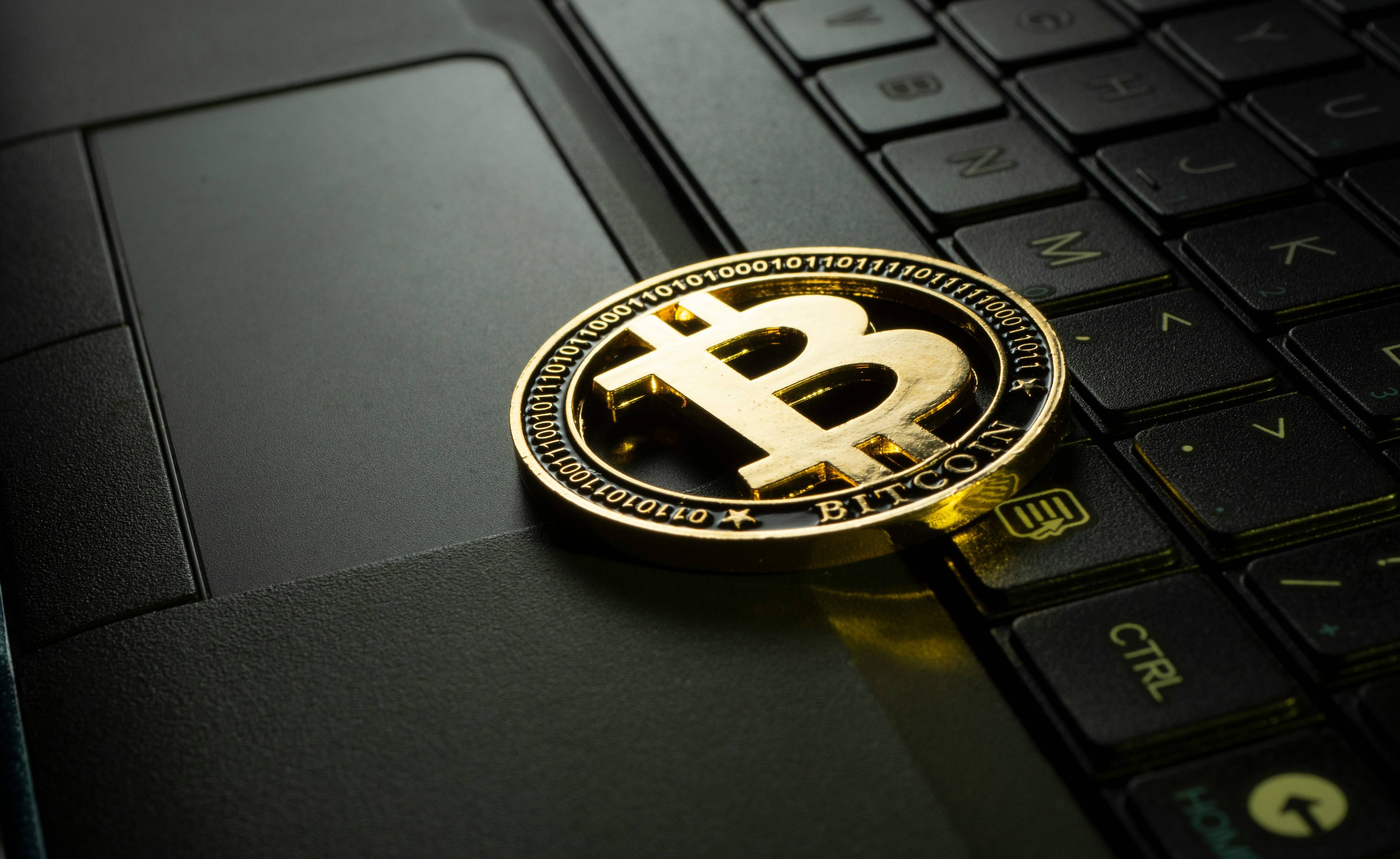 A gold Bitcoin coin rests on a laptop keyboard, partially covering the trackpad in dim lighting.