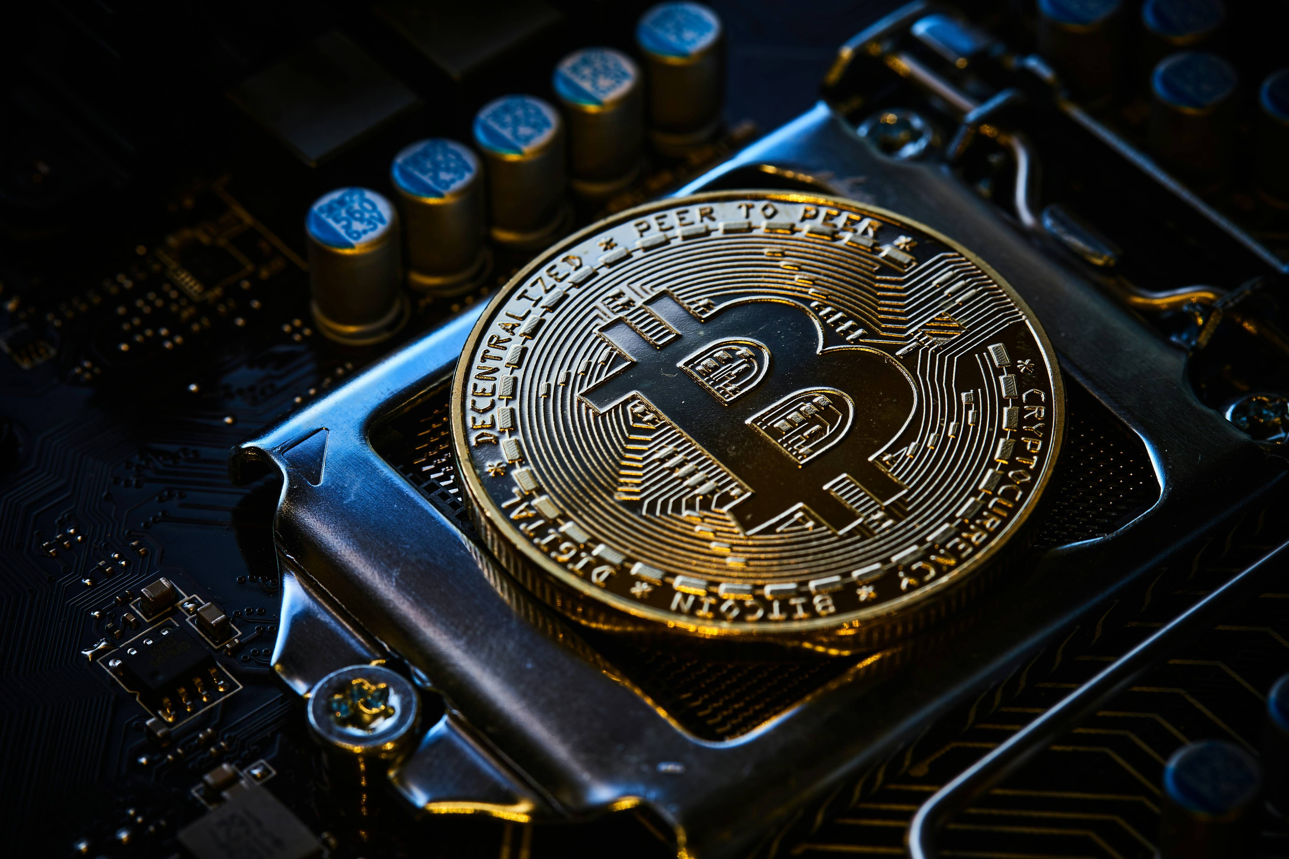A gold bitcoin rests on a computer motherboard with visible circuit details and text reads: digital, decentralized, peer to peer.