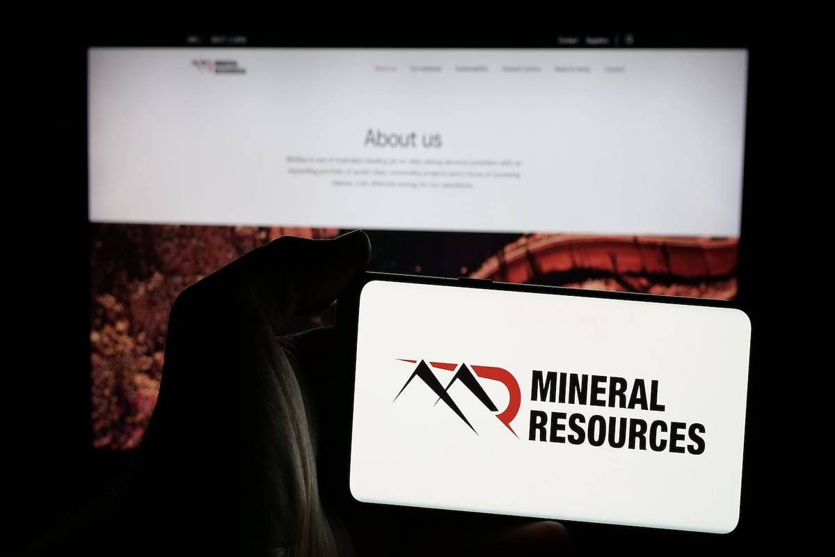 A hand holds a phone displaying "MINERAL RESOURCES" logo, in front of a computer screen showing an "About us" page.
