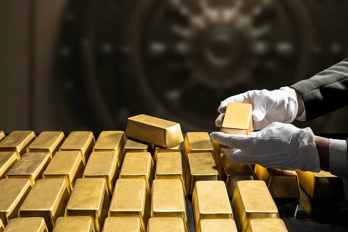 A pair of white-gloved hands holds a gold bar over a stack of gold bars with a vault in the background.