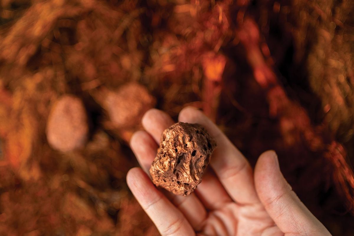 A person's hand holds a piece of copper metal ore above a blurry copper-colored background.