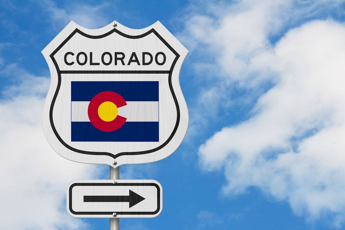 A road sign displaying the Colorado flag and an arrow, set against a blue sky with white clouds.
