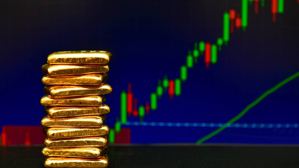 A stack of gold bars on a table in front of a rising stock market chart.