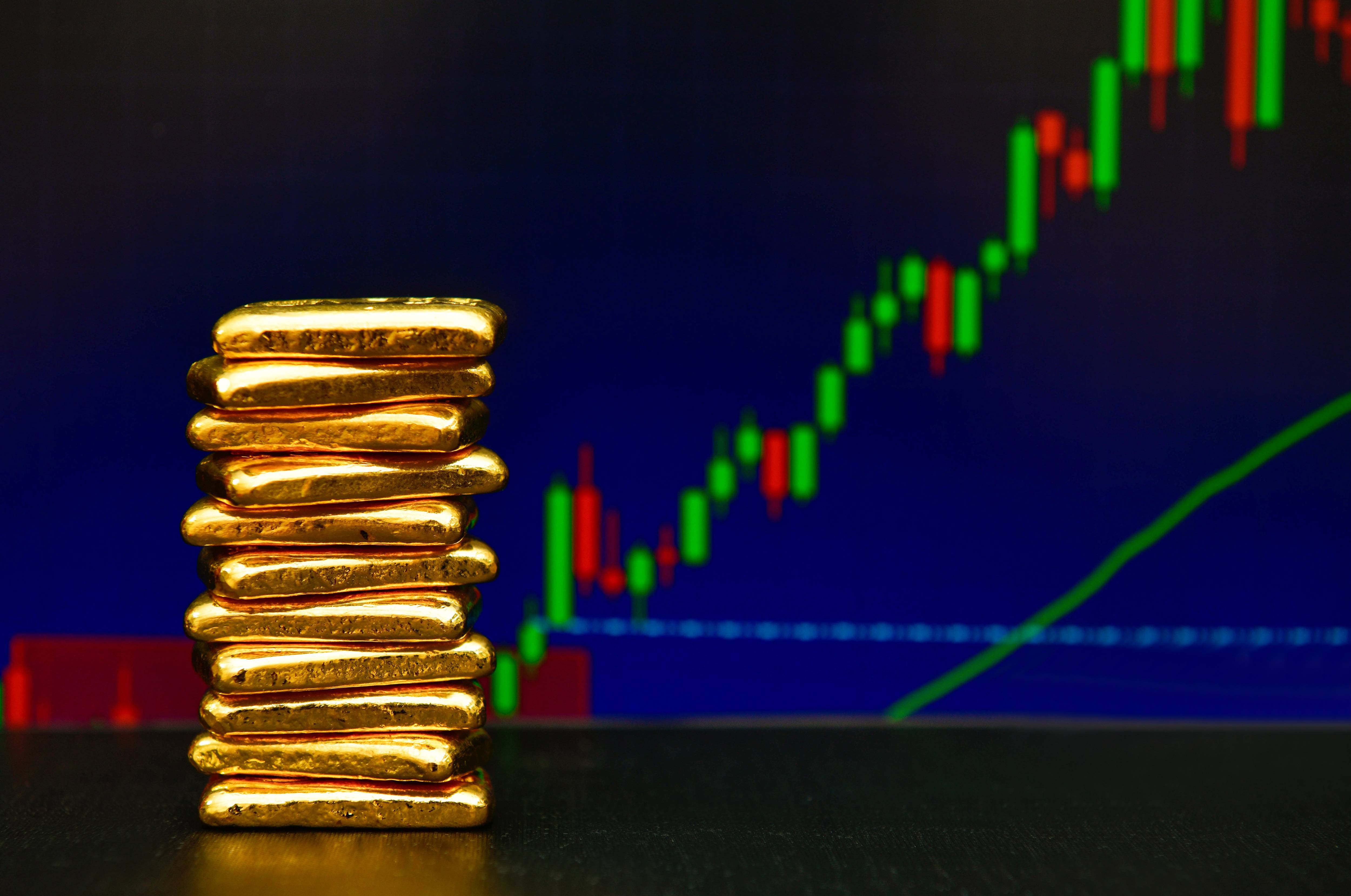 A stack of gold bars on a table in front of a rising stock market chart.