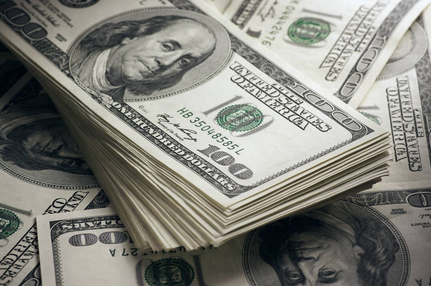 A stack of US hundred-dollar bills lies on top of more scattered banknotes.