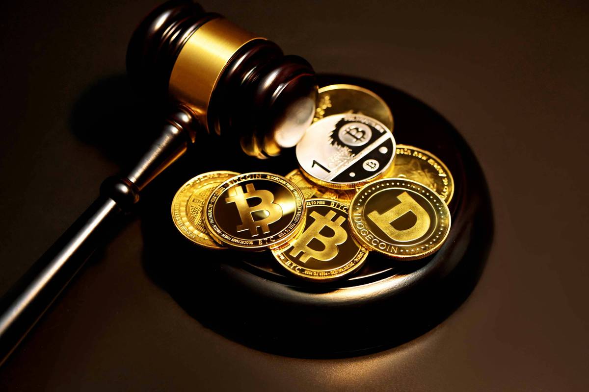 A wooden gavel rests on a pile of gold-colored cryptocurrency coins with Bitcoin and Dogecoin logos.