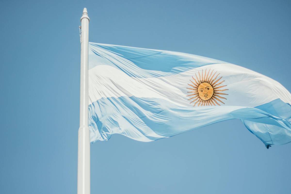 Argentinian flag with sun emblem waves on a white pole against a clear blue sky.