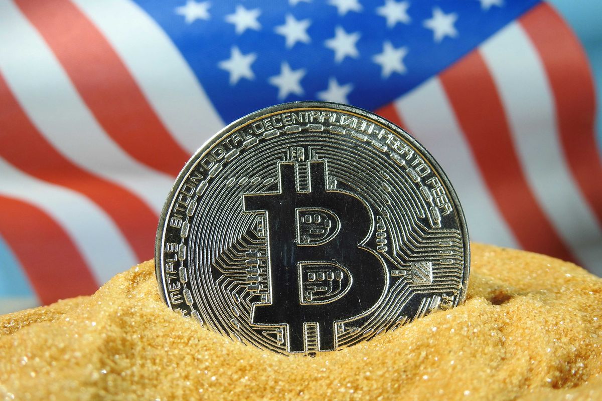 Bitcoin coin in sand with blurred US flag in background.