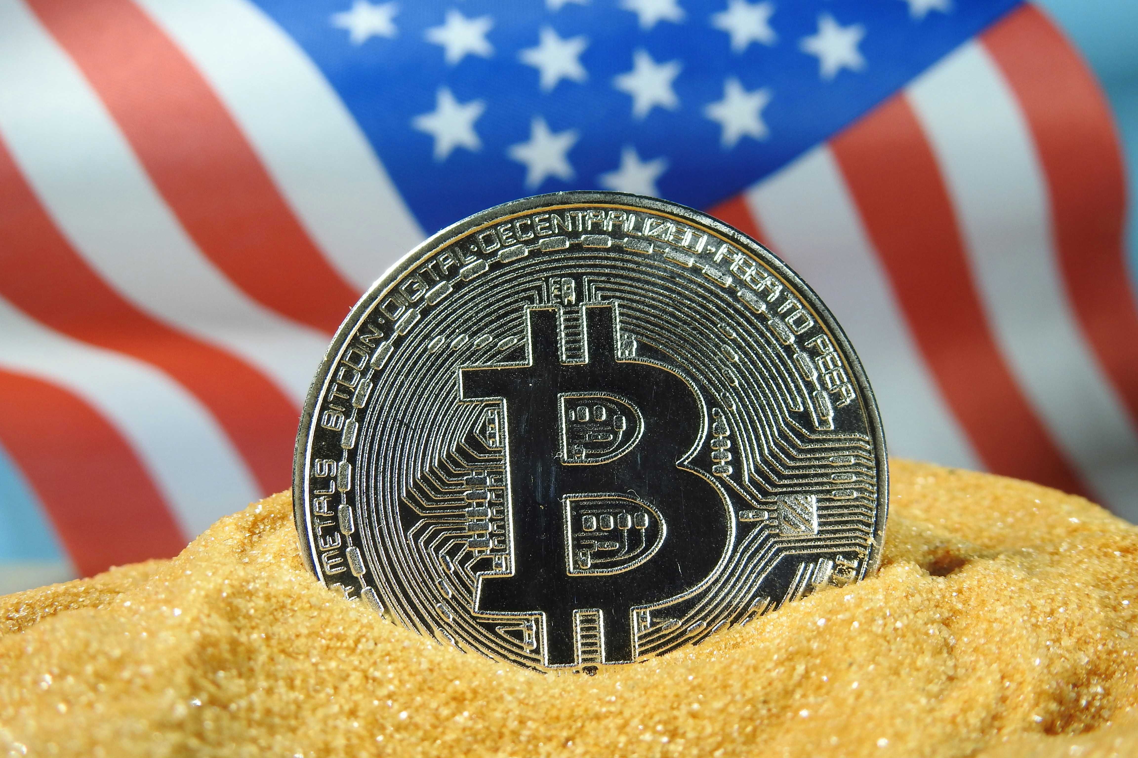 Bitcoin coin in sand with blurred USA flag in background.