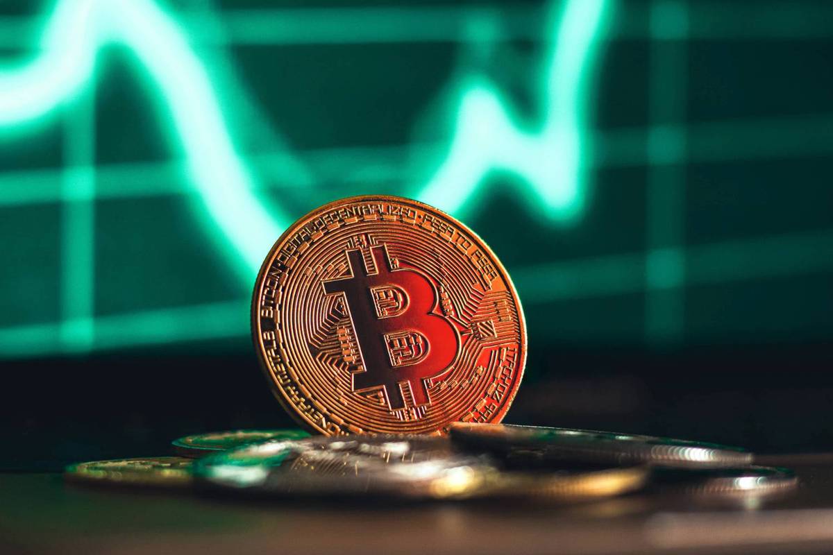 Bitcoin coin stands upright on pile of coins with green line graph in the background.
