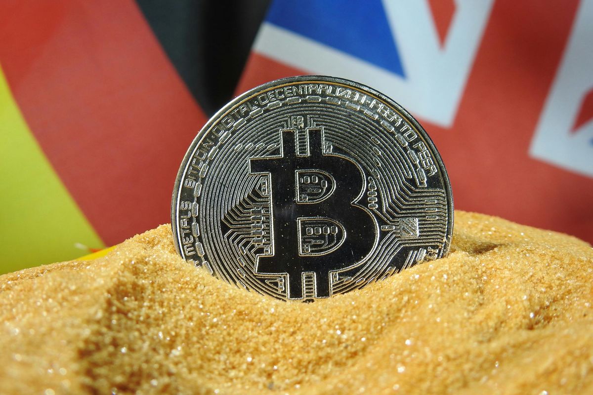 Bitcoin partially buried in sand, with a blurred colorful flag background.