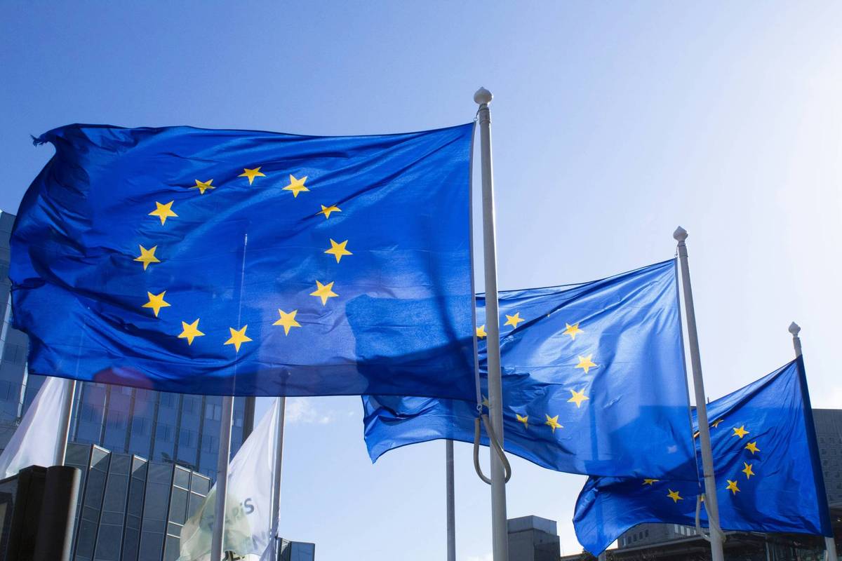 Blue flags with yellow stars wave in front of modern glass buildings under a clear blue sky.