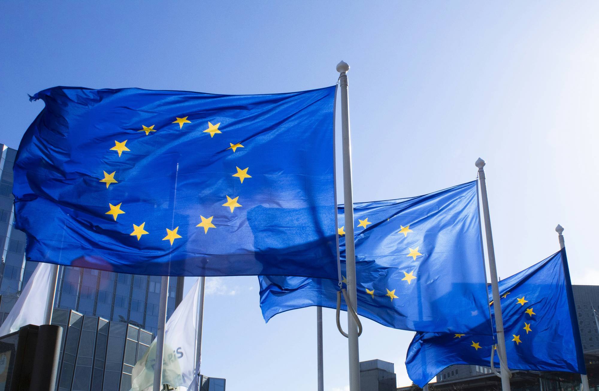 Blue flags with yellow stars wave in front of modern glass buildings under a clear blue sky.