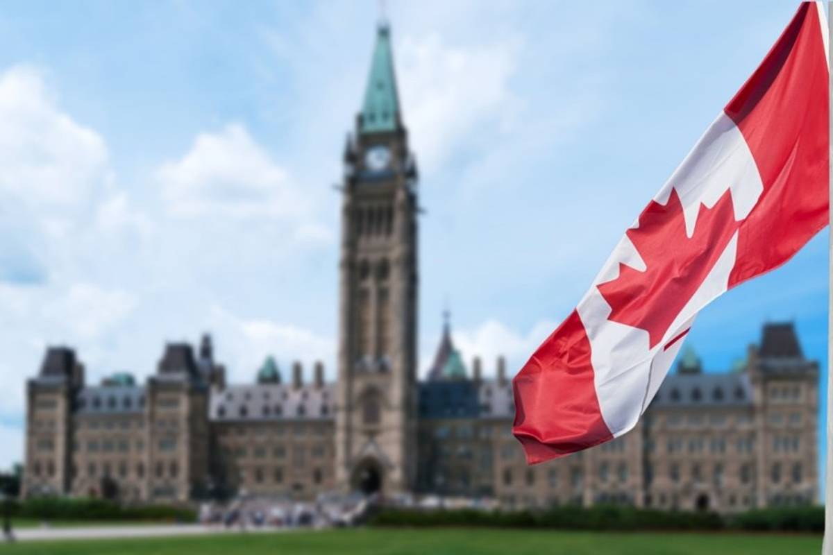 Canadian flag waves in the foreground with parliament and cloudy sky in the background.