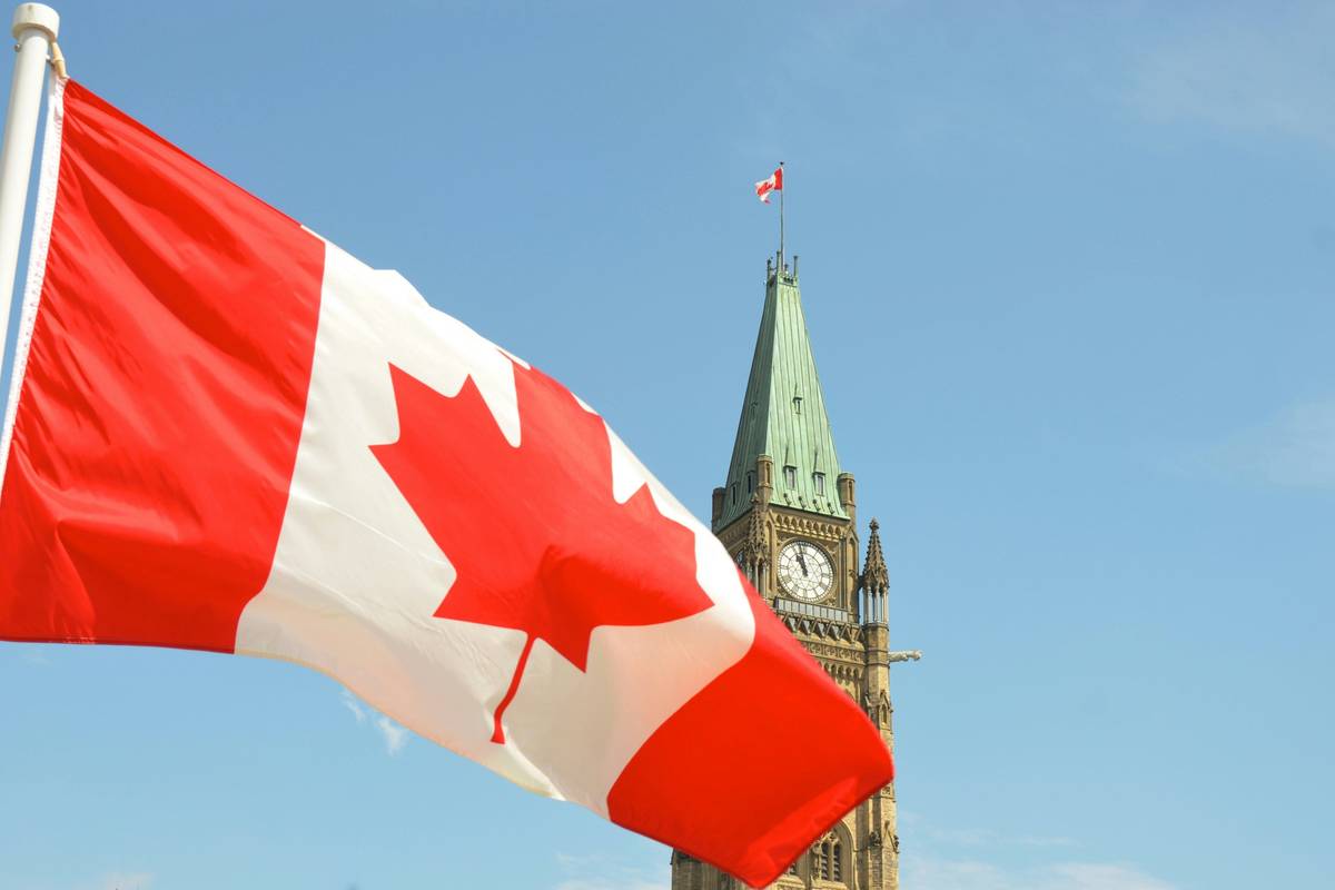 Canadian flag waving near Parliament clock tower against a clear blue sky.