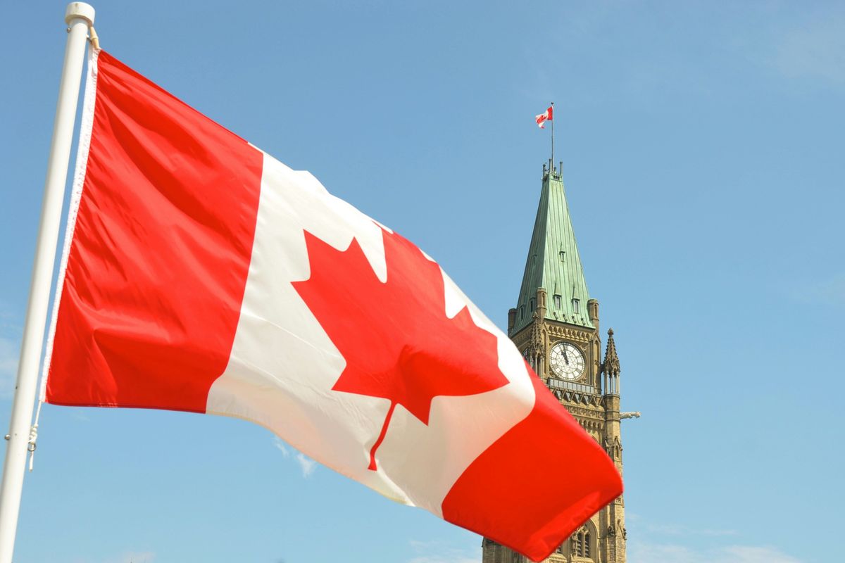 Canadian flag waving near Parliament clock tower against a clear blue sky.