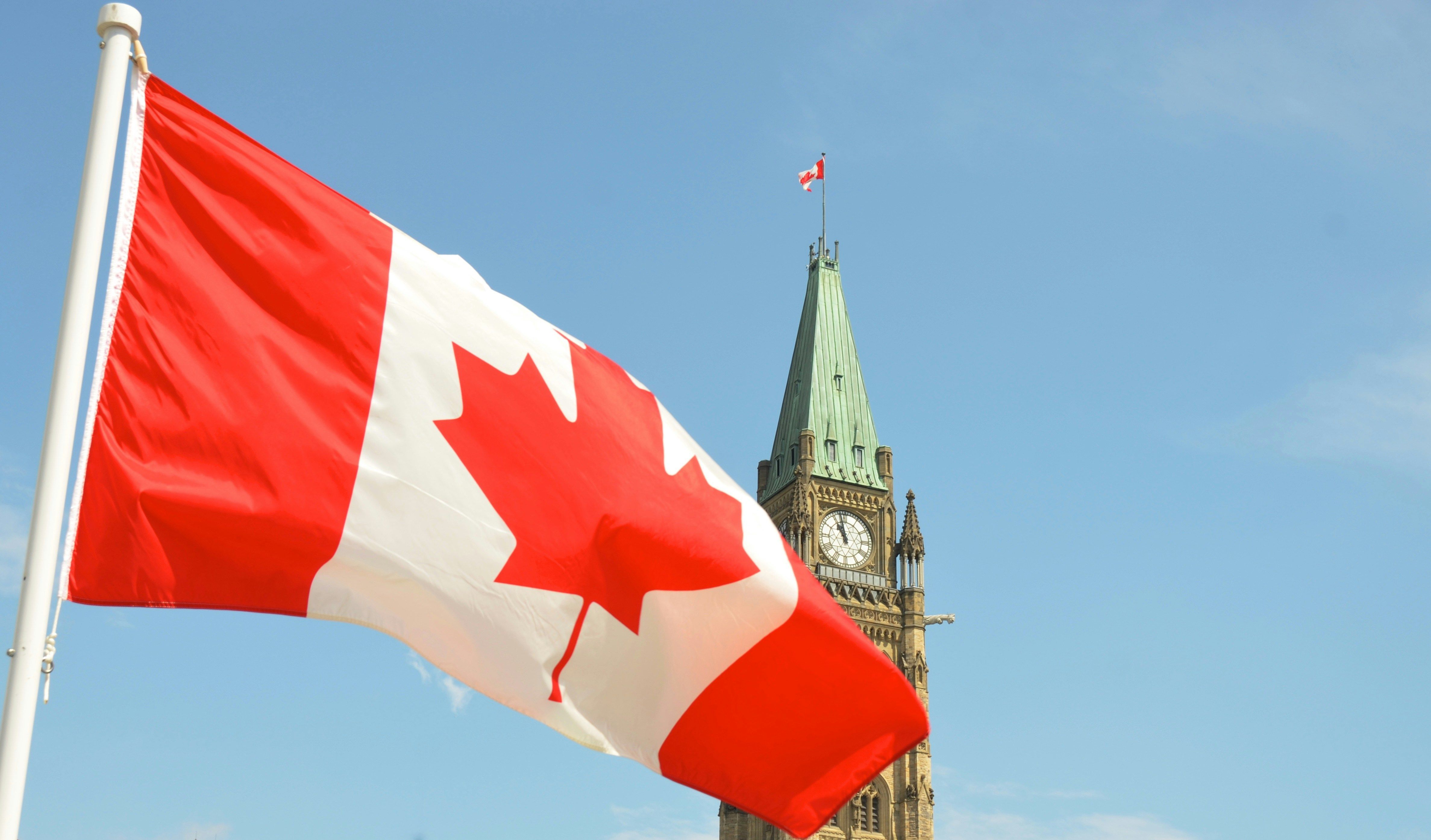 Canadian flag waving near Parliament clock tower against a clear blue sky.
