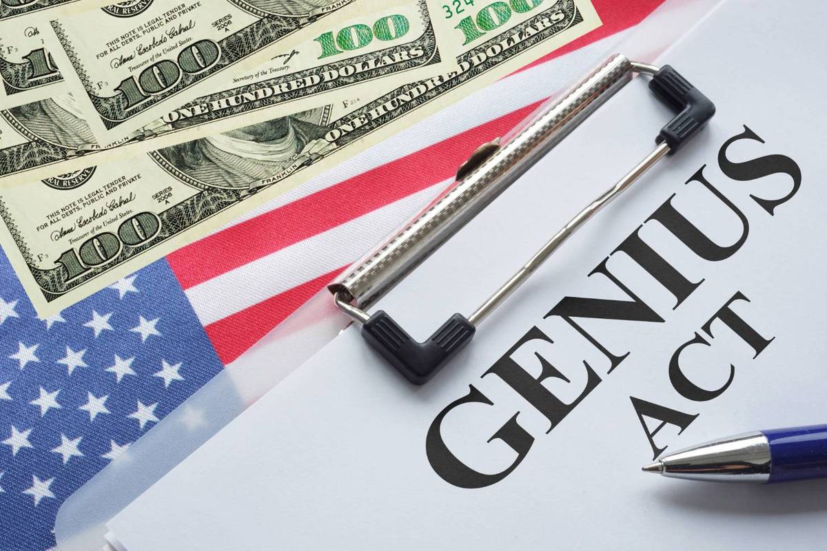 Clipboard with paper labeled "GENIUS Act" overlaid with US$100 bills on an American flag background.