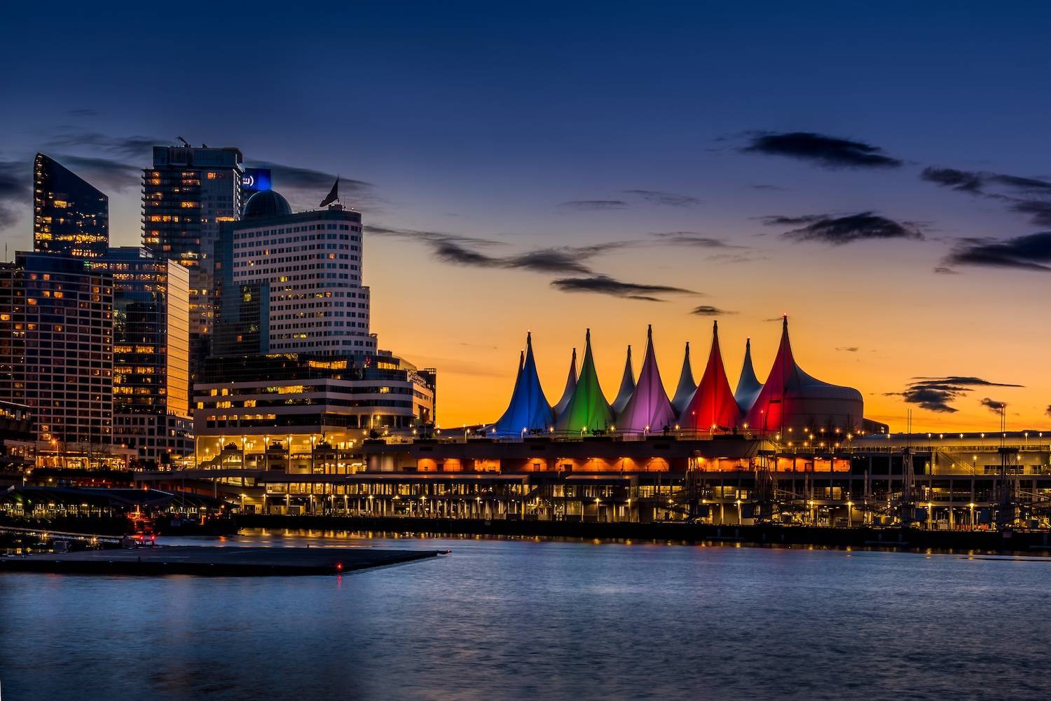 Colorful sails illuminate a waterfront building against a sunset sky, with skyscrapers on the left.