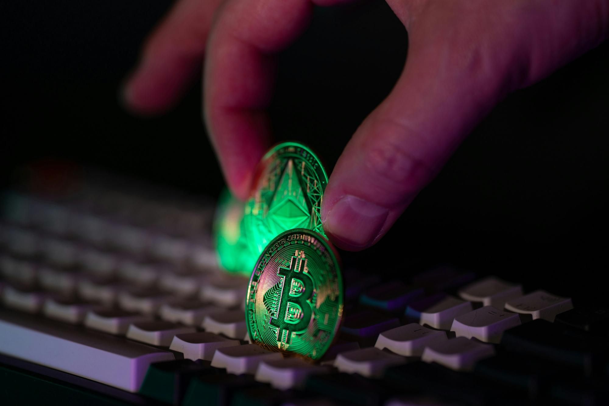 Fingers holding Bitcoin and Ether coins over a keyboard, illuminated by green light.