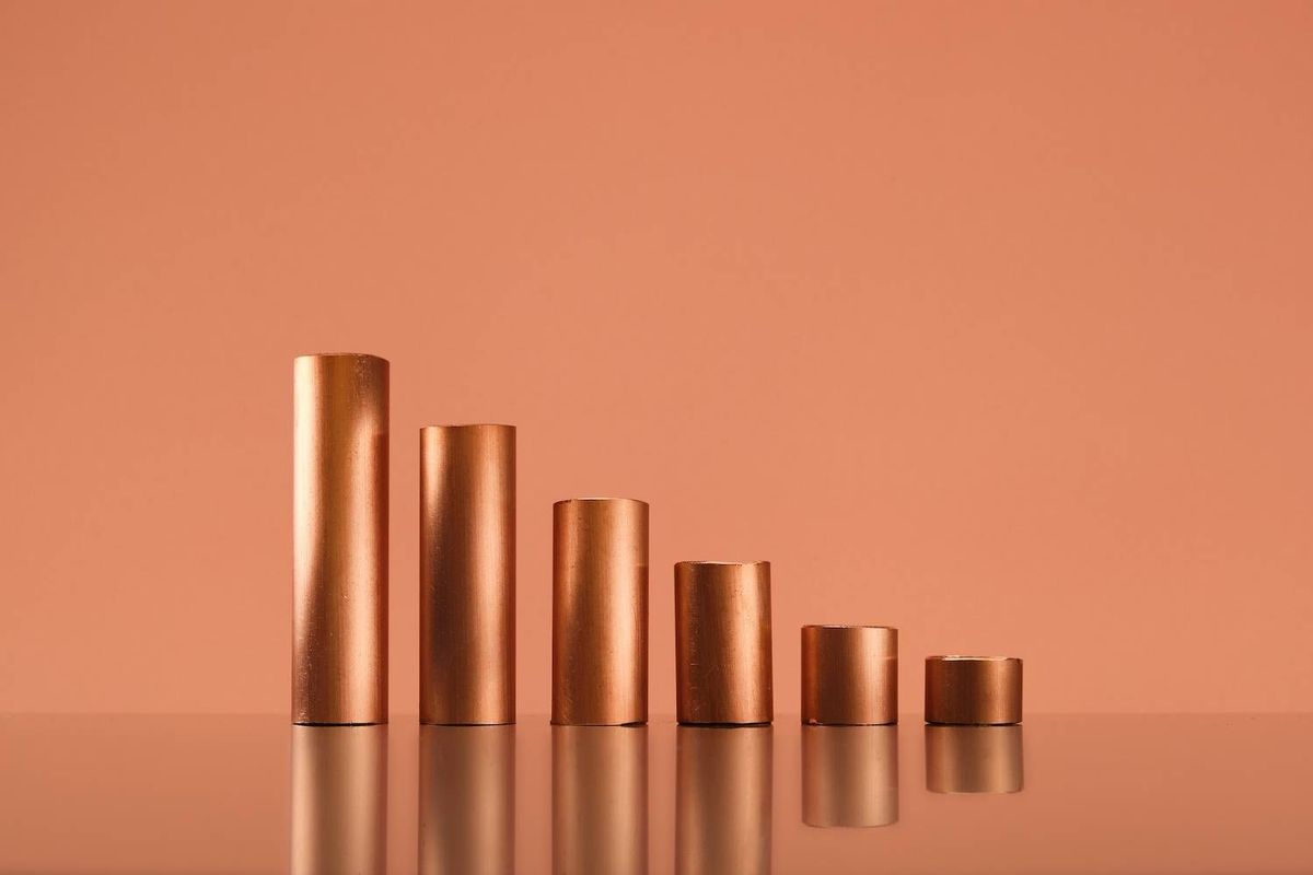 Five bronze cylinders of varying heights stand on a reflective surface against an orange background.