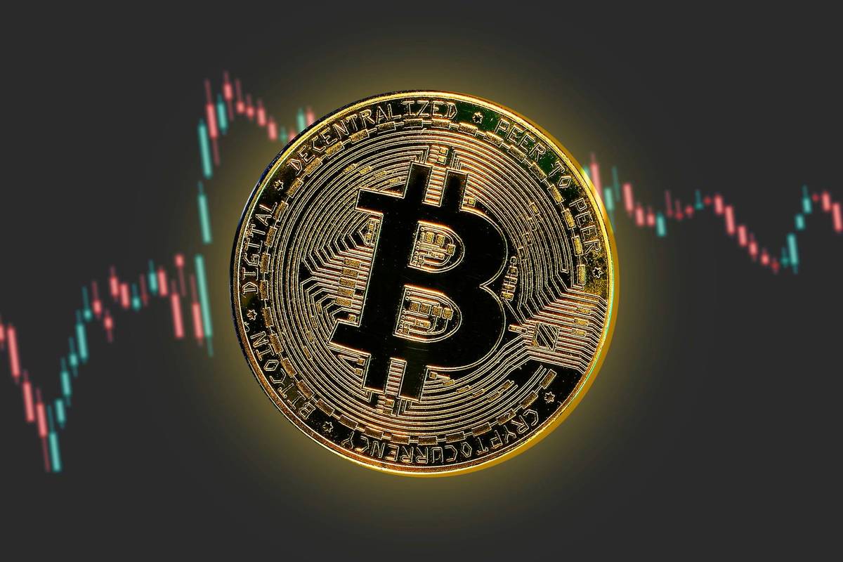 Glowing Bitcoin coin centered over gray background with fluctuating red and blue candlestick charts.