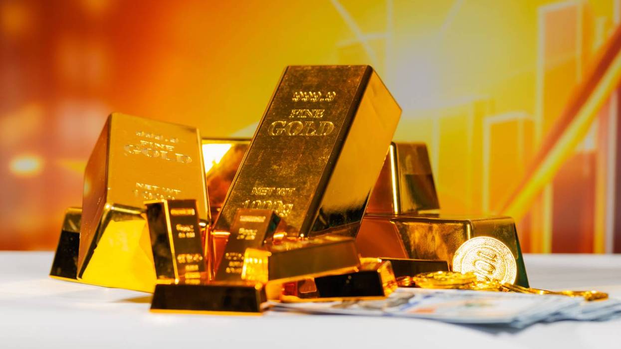 Gold bars, coins, and cash on a table against a golden-yellow background.