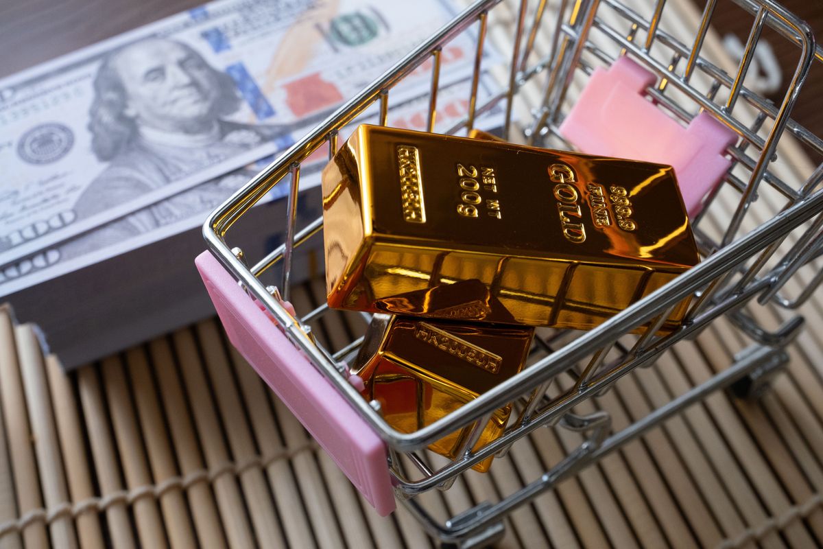 Gold bars in a small shopping cart beside a stack of US hundred-dollar bills.