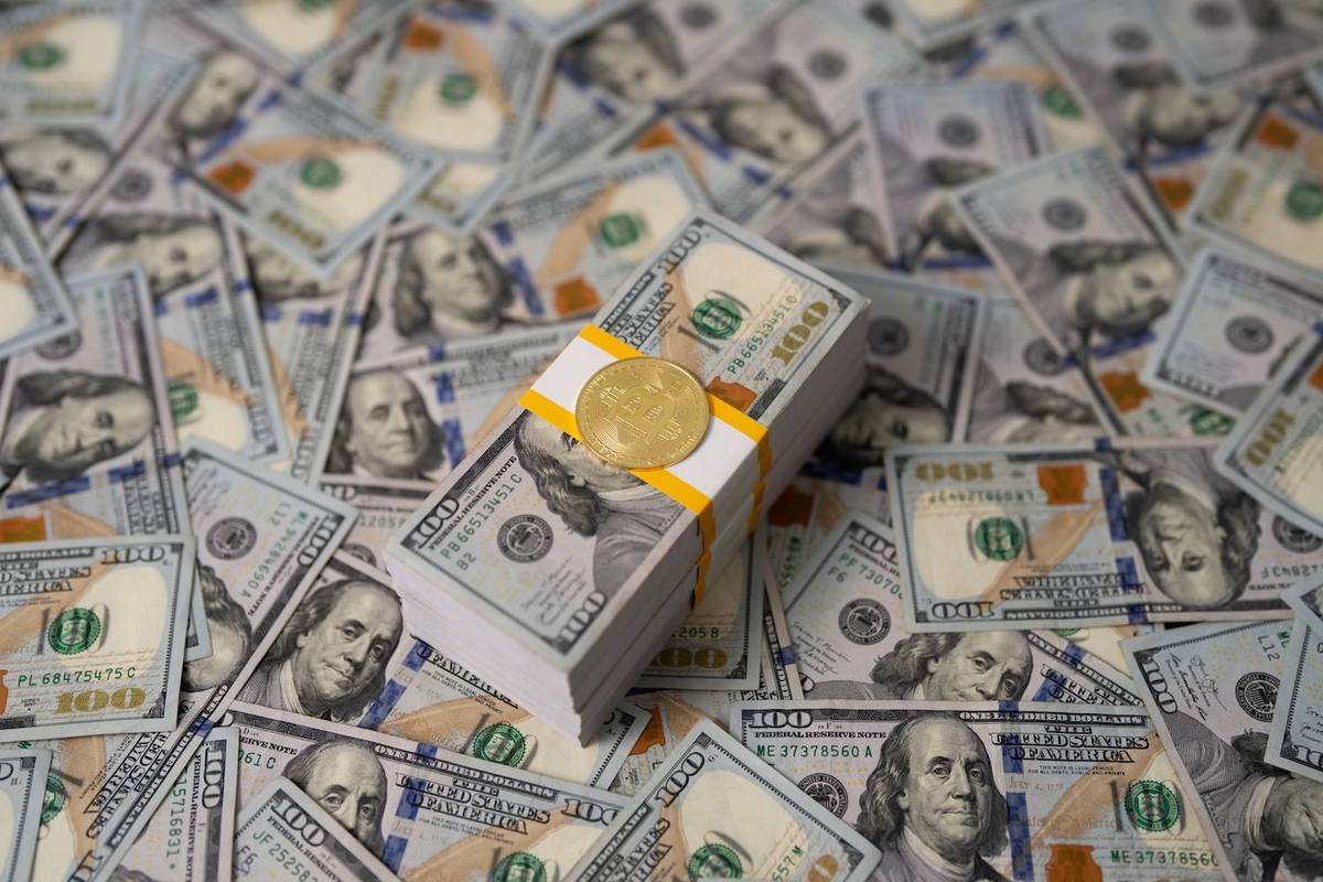 Gold Bitcoin on stack of 100-dollar bills among scattered banknotes.