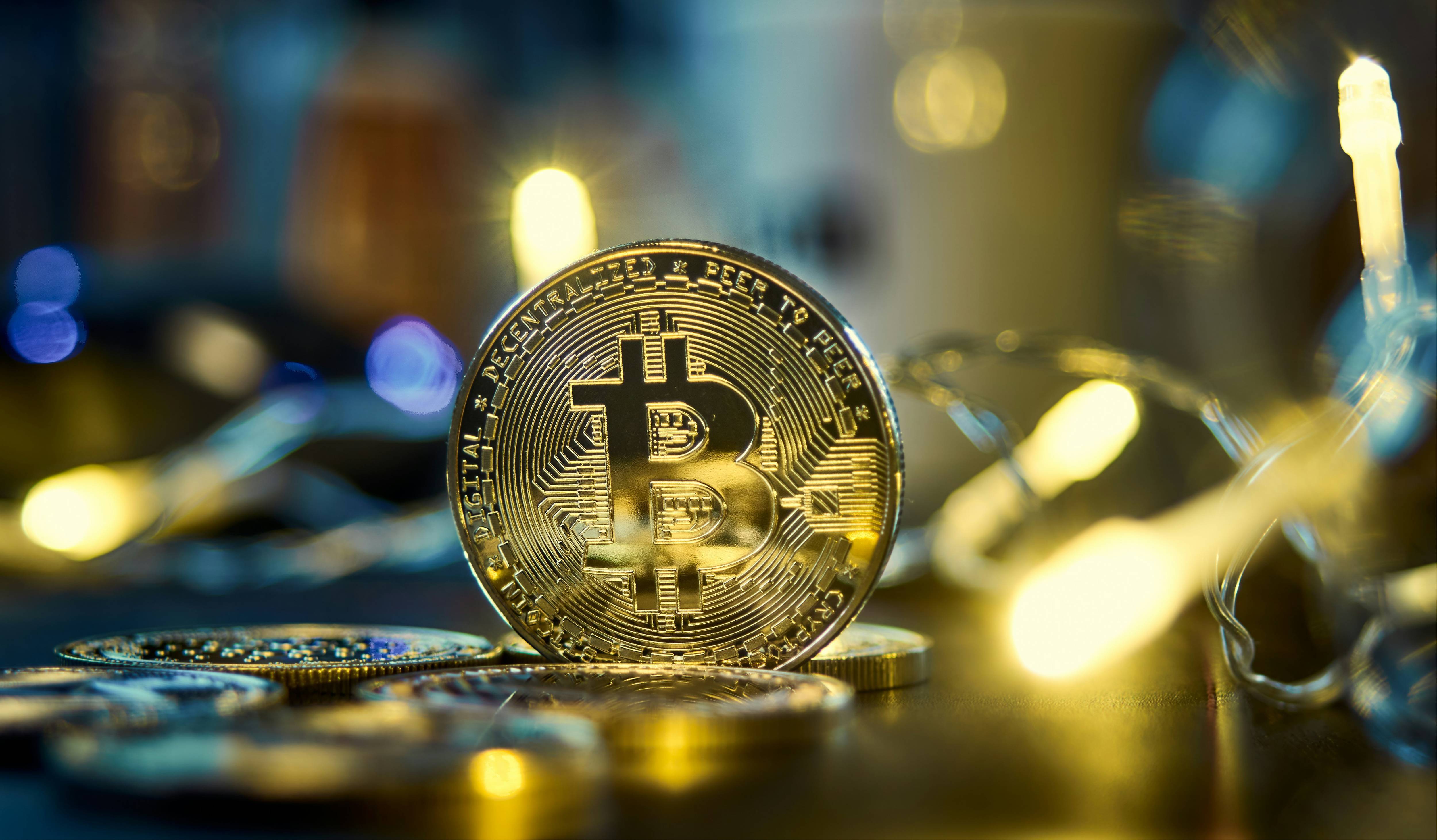 Golden Bitcoin coin stands upright among blurred fairy lights on a dark surface.