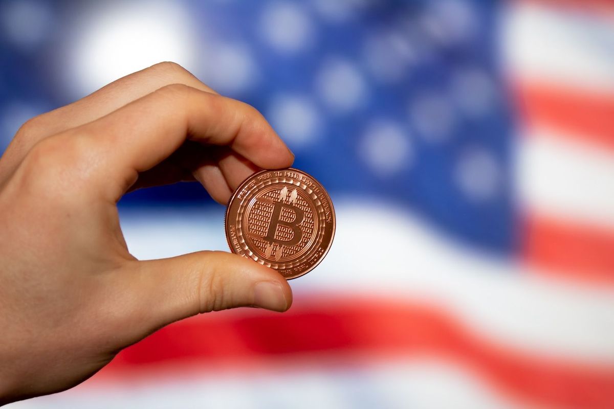 Hand holding a copper Bitcoin in front of US flag.