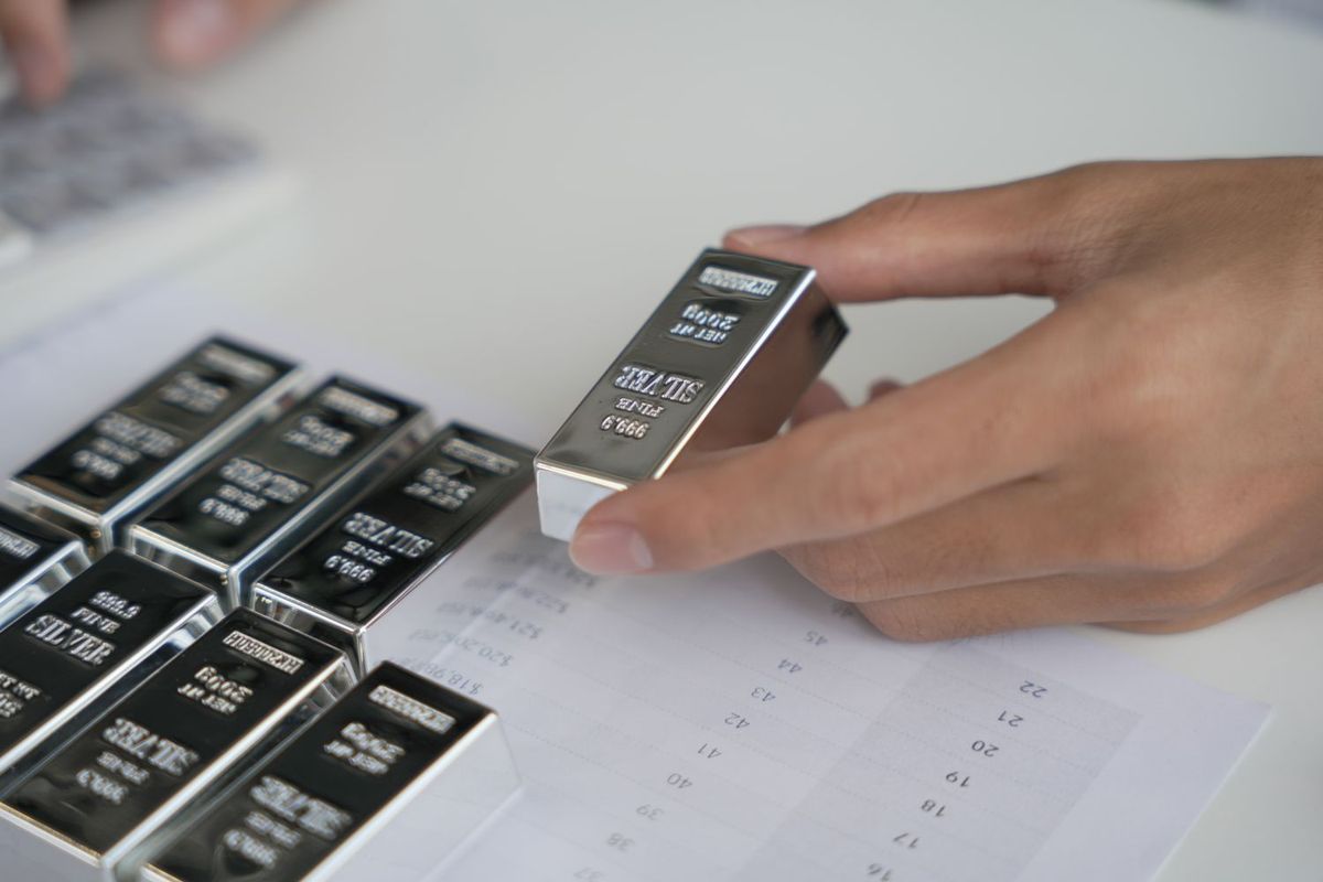 Hand holding a silver bar near a paper sheet with price charts and numbers; other silver bullion bars nearby.