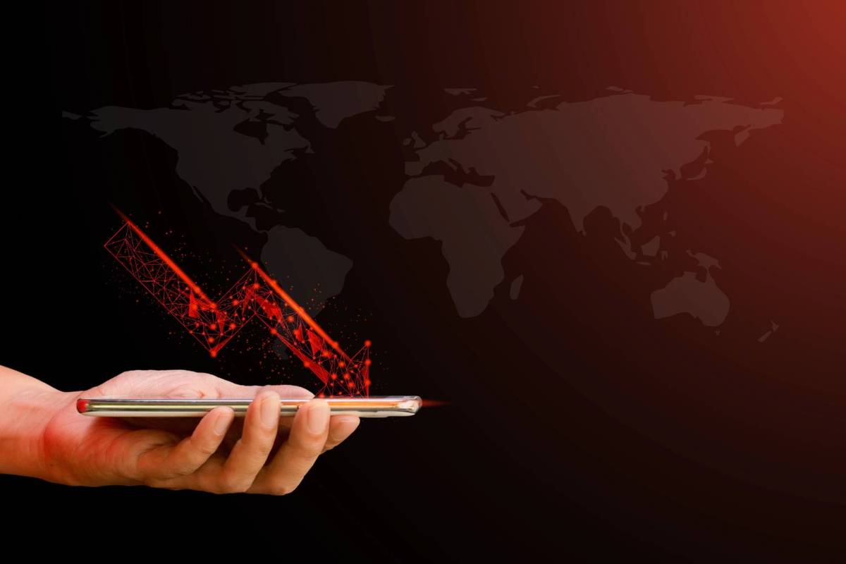 Hand holding a smartphone projecting a red downward graph, world map background.
