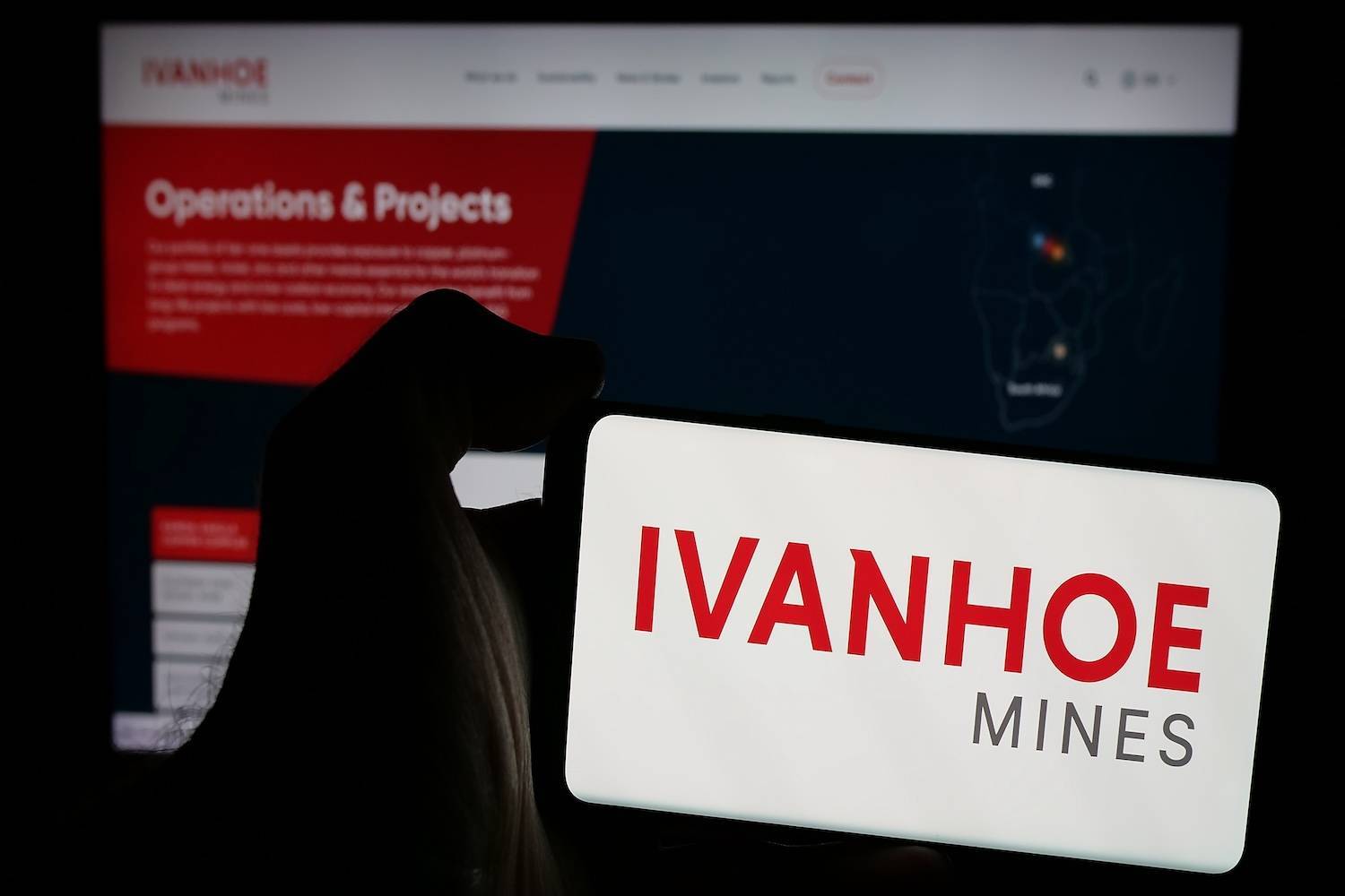 Hand holding phone with Ivanhoe Mines logo; website in the background.