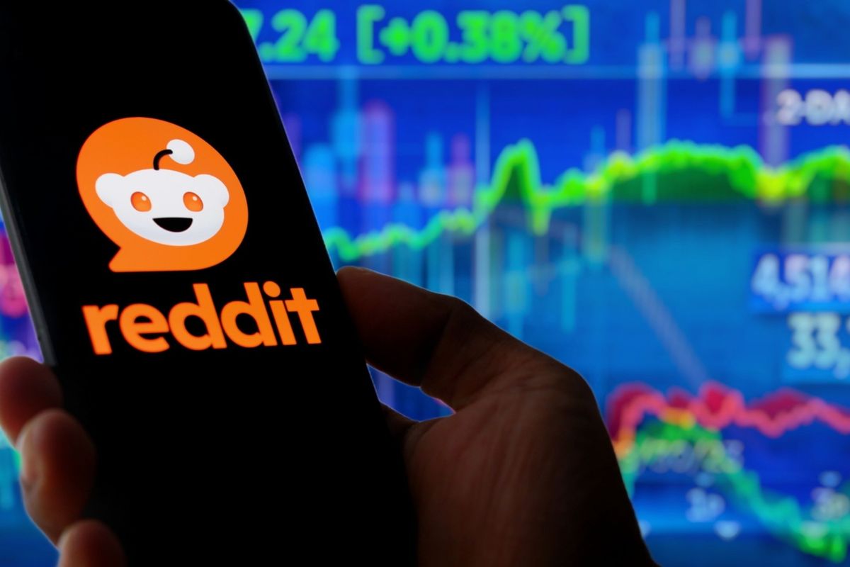 Hand holding smart phone with the Reddit logo in front of tech stock charts.