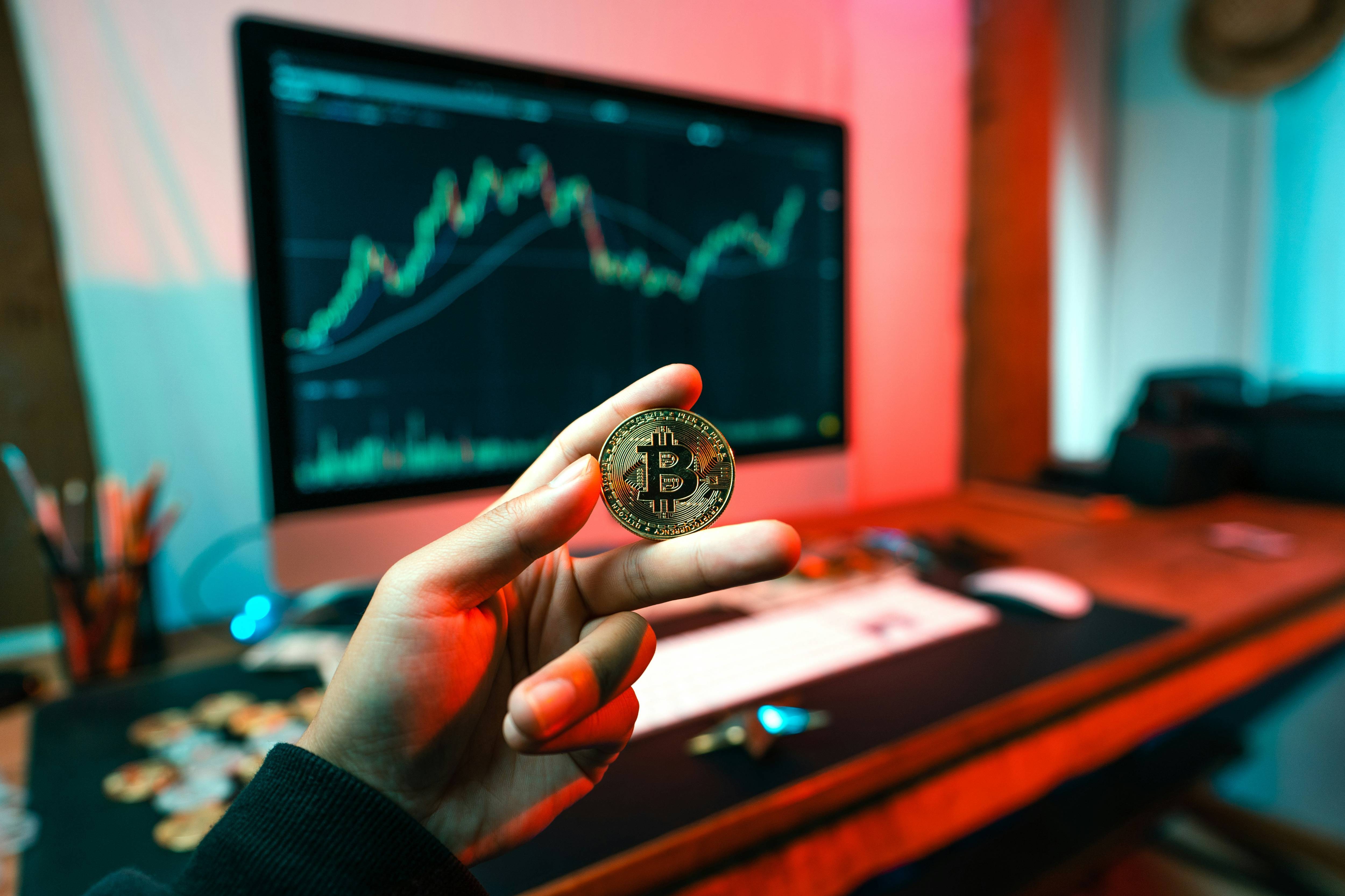 Hand holds Bitcoin in front of desk with computer displaying stock charts and graphs.