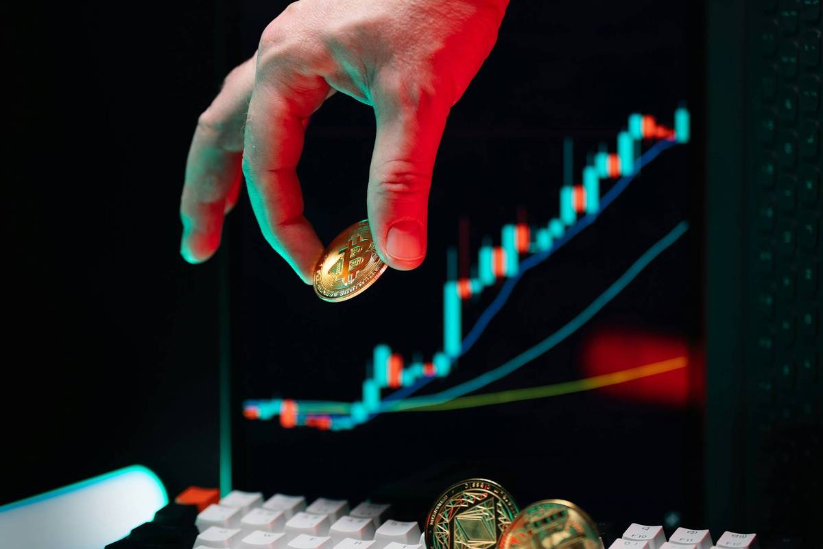 Hand holds Bitcoin over keyboard with rising cryptocurrency charts on a black screen.