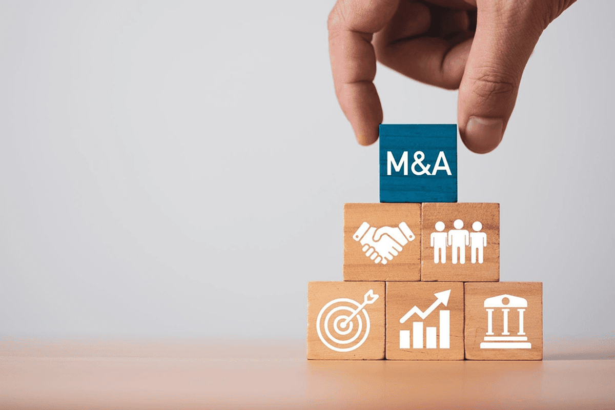 Hand places a blue block labeled "M&A" atop a pyramid of wooden blocks with business icons.