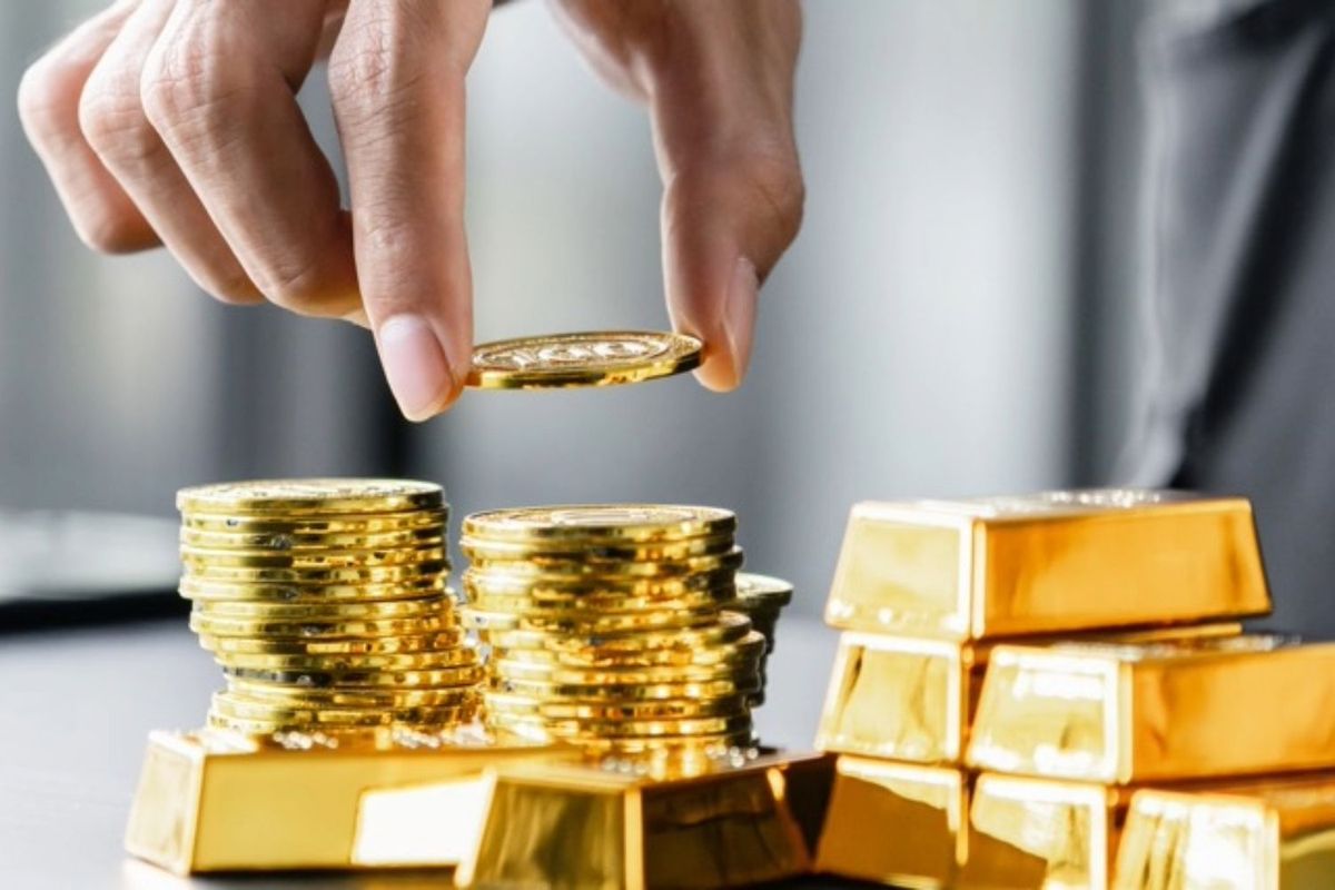 Hand stacking gold coins next to gold bars on a reflective surface.
