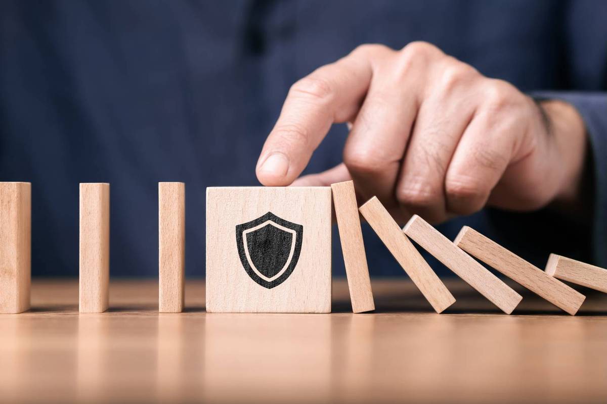Hand stops dominoes from falling using a block with a shield icon on a wooden surface.