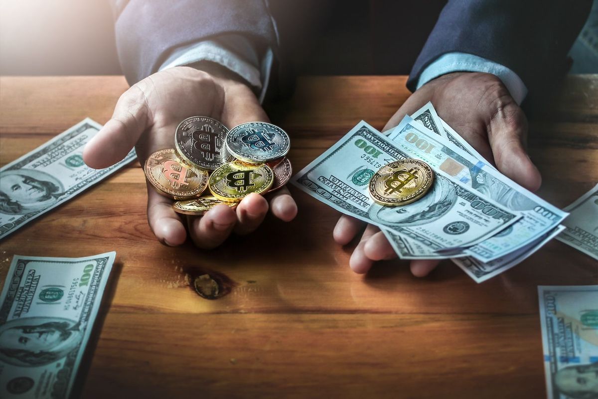Hands holding Bitcoin coins and US dollar bills on a wooden table.