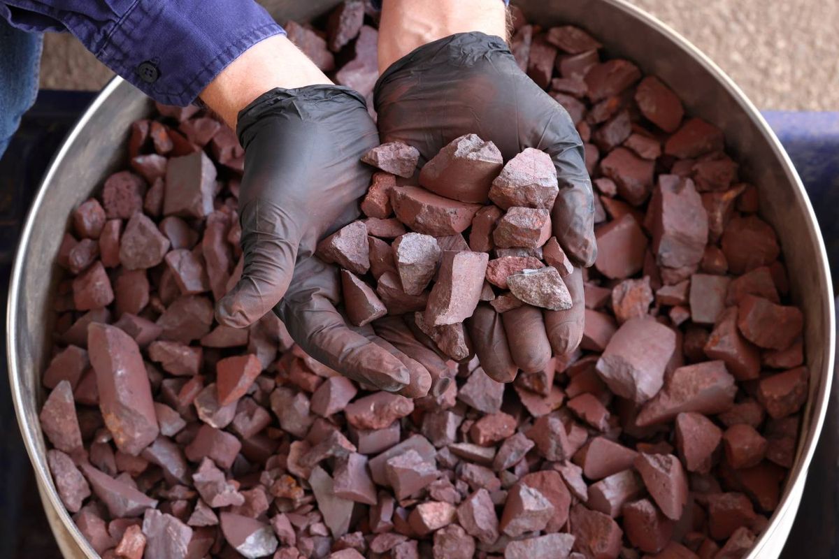 Hands in black gloves holding reddish-brown iron ore pieces from Pilbara over a metal container filled with iron ore.
