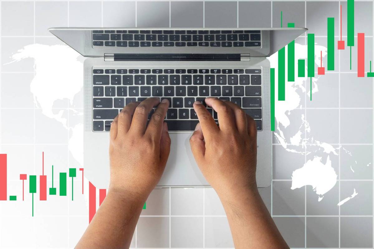 Hands typing on laptop keyboard with world map and red/green stock charts in the background.