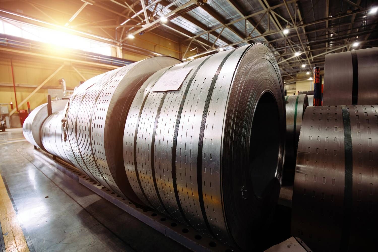 Large rolls of sheet metal in a factory warehouse with bright lighting.